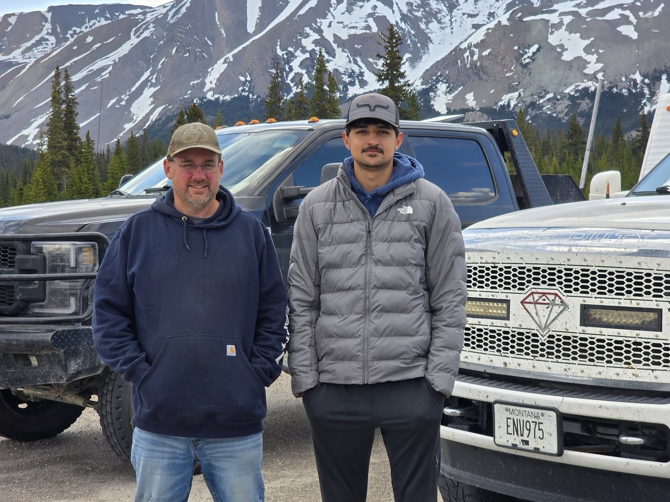 Two crew members standing beside a truck with snowy mountains behind them.